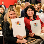 Dwie kobiety siedzą wśród innych uczestników i trzymają listy gratulacyjne.