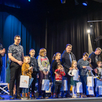 Na scenie stoi grupa dzieci, trzymają białe torby z logo Białegostoku, na scenie stoją również opiekunowie, zastępca prezydenta Rafał Rudnicki i duchowny.