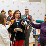 Kobieta udziela wywiadu podczas konferencji, obok stoją dziennikarze z mikrofonami.