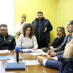 Uczestnicy spotkania siedzą przy stole i stoją dookoła sali.