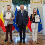 Dwoje uczniów trzyma listy gratulacyjne i niewielki białe torebki z upominkami, po środku stoi prezydent Tadeusz Truskolaski.