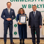 Kobieta trzyma dyplom, obok stoi dwóch mężczyzn.