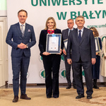 Kobieta trzyma dyplom, obok stoi dwóch mężczyzn, w tle stoją inni uczestnicy.