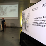 Zastępca prezydenta Rafał Rudnicki przemawia do mikrofonu, na pierwszym planie jest ekran z grafiką i tekstem „Inauguracja Roku Jubileuszowego Wydziału Architektury Politechniki Białostockiej”.