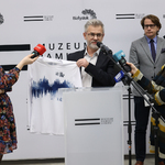 Prezes Fundacji Białystok Biega Grzegorz Kuczyński mówi do mikrofonów podczas konferencji, trzyma w rękach i pokazuje koszulkę.