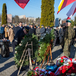 Uczestnicy uroczystości stoją przed pomnikiem pod którym leżą kwiaty.