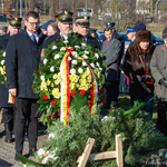 Zastępca prezydenta Tomasz Klim stoi za mundurowym, który trzyma wieniec, w tle inni uczestnicy wydarzenia.