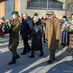 Kilku uczestników uroczystości idzie za żołnierzem z kwiatami w rękach.