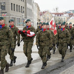 Żołnierze idą w szeregach przez Rynek Kościuszki, jeden z żołnierzy niesie flagę Polski.
