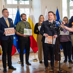 Przedstawiciel „Organizacji Pożytku Publicznego” stoi przed mikrofonami, obok stoją dziennikarze oraz inni uczestnicy.