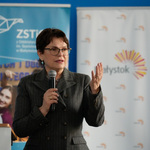 Kobieta mówi do mikrofonu, trzyma rękę uniesioną.