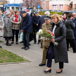 Uczestnicy wydarzenia tłoczno stoją na chodniku, kobieta niesie bukiet kwiatów, obok idzie mężczyzna, w tle zabudowania przy ul. Sienkiewicza.