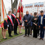 Poczet sztandarowy szkoły, obok stoi kilku uczestników wydarzenia, w tle zabudowania.