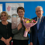 Troje uczestników stoi obok siebie, kobieta trzyma ulotkę oraz bukiet kwiatów.