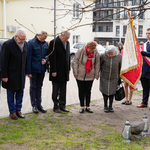 Kilkoro uczestniów stoi z pochyloną głową, obok stoi uczeń ze sztandarem, w tle zabudowania.