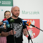 Marszałek Łukasz Prokorym udziela wywiadu podczas konferencji prasowej, obok dziennikarze trzymają mikrofony.