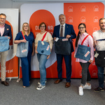 Sześcioro uczestników konferencji prasowej stoi obok siebie, każdy z nich trzyma torbę.