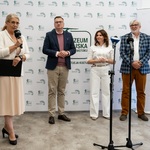 Troje uczestników konferencji stoi, obok prowadząca mówi do mikrofonu.