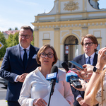 Kobieta mówi do mikrofonów podczas konferencji prasowej, obok stoją inni uczestnicy, w tle Brama Wielka.