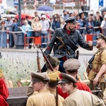 Grupa aktorów stoi na placu, w tle widownia.