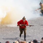 Aktor biegnie z bronią, w tle tuman kurzu.