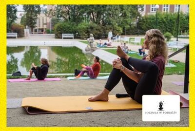 Kobiety siedzą na matach podczas treningu jogi w parku Różanka, w tle staw oraz rzeźba „Praczki”, w rogu zdjęcia logo Joginka w podróży.