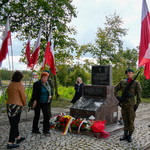 Uczestnicy uroczystości stoją przed pomnikiem pod którym stoją kwiaty.