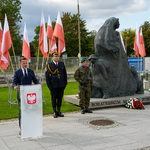 Przy pomniku „Matki Sybiraczki” stoją mundurowi, pod pomnikiem leżą wieńce i bukiety, w tle ustawione są stojaki z flagami Polski.