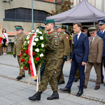 Wojewoda Jacek Brzozowski z asystą mundurowych idą z wieńcem w trakcie uroczystości.
