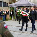 Mężczyzna w asyście funkcjonariusza Straży Miejskiej niosącego wieniec idzie podczas uroczystości.