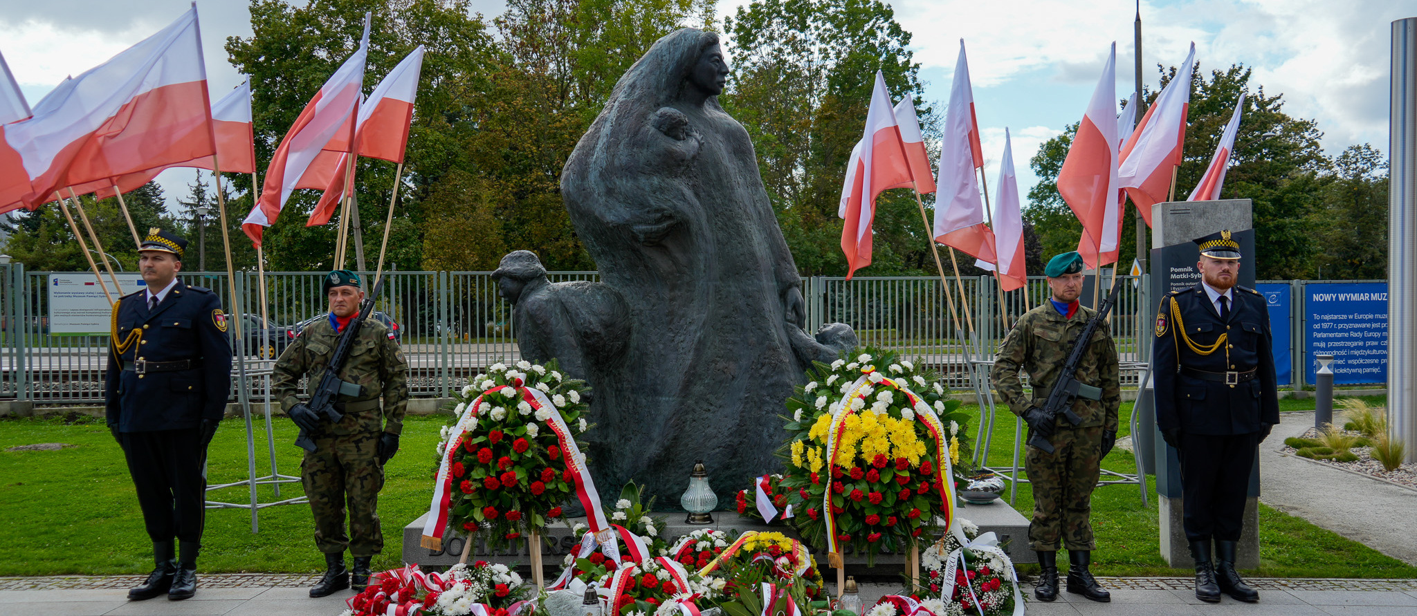 Przy pomniku „Matki Sybiraczki” stoją mundurowi, pod pomnikiem leżą wieńce i bukiety, w tle ustawione są stojaki z flagami Polski.