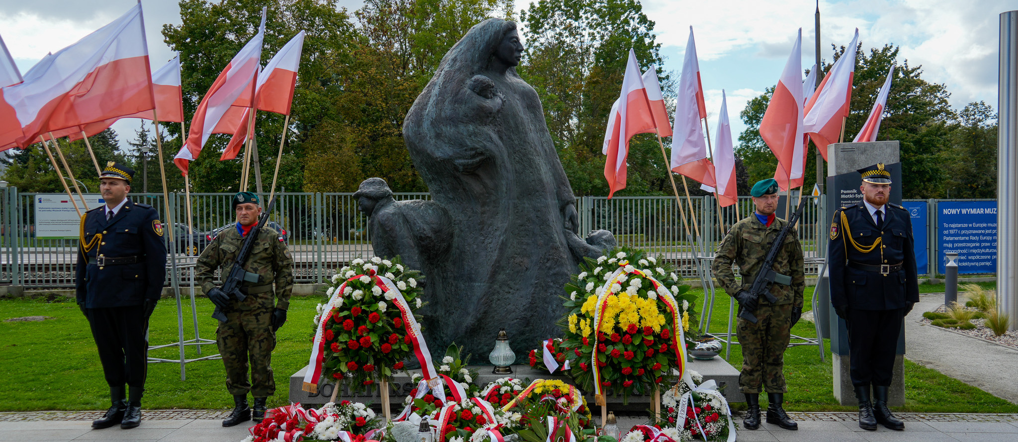Przy pomniku „Matki Sybiraczki” stoją mundurowi, pod pomnikiem leżą wieńce i bukiety, w tle ustawione są stojaki z flagami Polski.