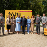 Uczestnicy konferencji stoją przed żółtą ścianka, obok stoi żółty fotel.