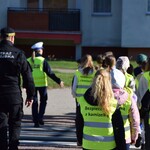 Dzieci w kamizelkach odblaskowych przechodzą przez przejście dla pieszych, przy nich strażnik miejski i policjantka