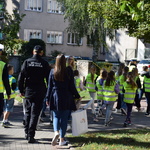 Dzieci w kamizelkach odblaskowych idą chodnikiem, za nimi strażnik miejski i policjantka