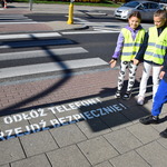 Dzieci w kamizelkach odblaskowych wskazują napis na chodniku 