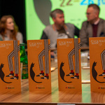 Ulotki promujące festiwal „Ciekawi Świata” stoją na stole, w tle siedzą uczestnicy konferencji.