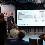 Uczestnicy konferencji stoją zwróceni w stronę ekranu na którym wyświetlany jest tekst.