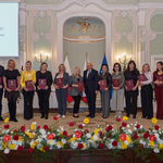 Kilkanaście wyróżnionych osób stoi z teczkami podczas uroczystości.
