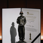 Statuetka nagrody BohaterON oraz dyplom. Statuetka przedstawia postać w mundurze, trzymającą dokument. Dyplom leży otwarty obok.