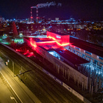 Widok na nowoczesny budynek muzeum oświetlony na czerwono, w tle widać dymiące kominy i miasto nocą.