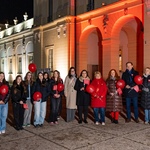 Grupa osób stoi na zewnątrz w nocy, trzymając czerwone balony i transparenty. W tle widoczna jest oświetlony Pałac Branickich.