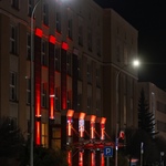 Obrazek przedstawia budynek oświetlony czerwonymi światłami w nocy. Na pierwszym planie widać zaparkowane samochody oraz znaki drogowe.
