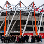 Pod budynkiem Chorten Areny stoi grupa osób uczestniczących w konferencji prasowej.
