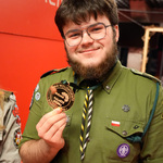 Młody mężczyzna w mundurze harcerskim trzyma medal. Uśmiecha się, a w tle widać czerwoną ścianę z napisem.