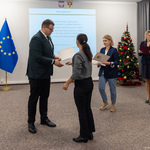 Na uroczystości wręczenia dyplomów, Zastępca prezydenta Tomasz Klim przekazuje dokument kobiecie. W tle widoczne flagi Polski i Unii Europejskiej oraz choinka.