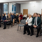 Na sali znajduje się grupa osób siedzących na krzesłach. W tle widać okna i zegar na ścianie.