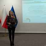Na zdjęciu stoi kobieta trzymająca dokumenty, w tle widoczne flagi Polski i Unii Europejskiej. Na ścianie wyświetlony tekst dotyczący ślubowania.
