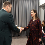 Zastępca prezydenta Tomasz Klim i kobieta podają sobie ręce w trakcie ceremonii. W tle siedzi kilka osób, a za nimi widać zasłony.