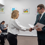 Zastępca prezydenta Tomasz Klim i nauczycielka podają sobie ręce podczas ceremonii wręczenia dokumentów. W tle widać kilka obrazków na ścianie oraz siedzące osoby.